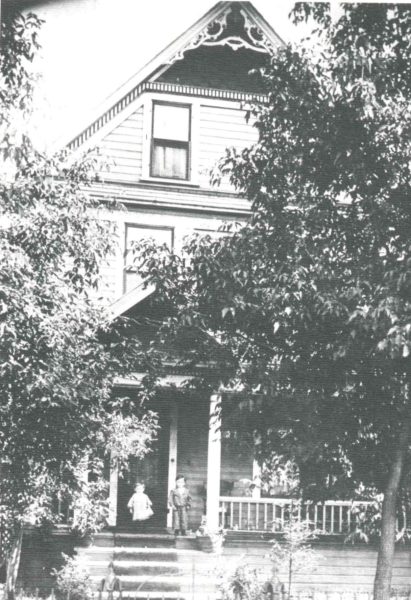 A grainy black and white photo shows a house partially obstructed by trees. Two children stand on the front porch.