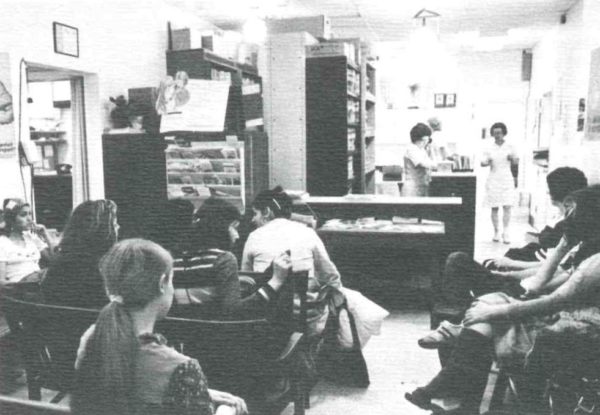 A grainy black and white photo shows the waiting room of the clinic filled with patients. Three medical staff are working in the background.
