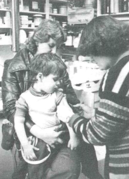 A grainy black and white photo shows a parent holding young child as they receive a vaccination in their arm from medical staff.