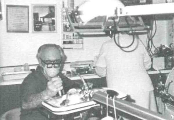 A grainy black and white photo shows two medical staff in the clinic during the 1970s. One wears a mask with thick glasses on and is operating a piece of medical equipment. The other has their back to the camera and is working at a counter.