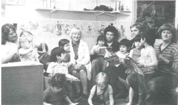A grainy, black and white photo from the 1970s of Anne Ross wearing a white lab coat and sitting with children from the daycare.