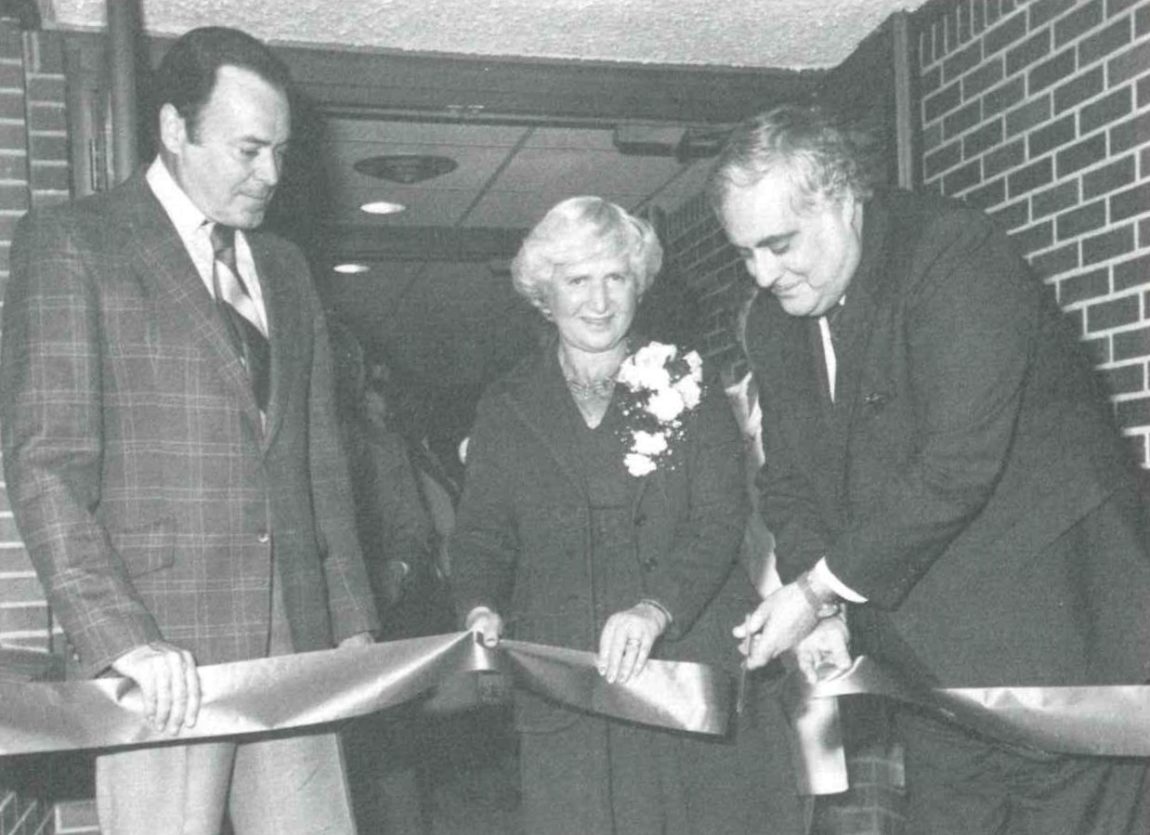 A grainy black and white photo shows Anne Ross standing alongside two politicians, holding a large ribbon outside the doors of Anne Ross Day Nursery. One of the politicians is cutting the ribbon with a pair of scissors.