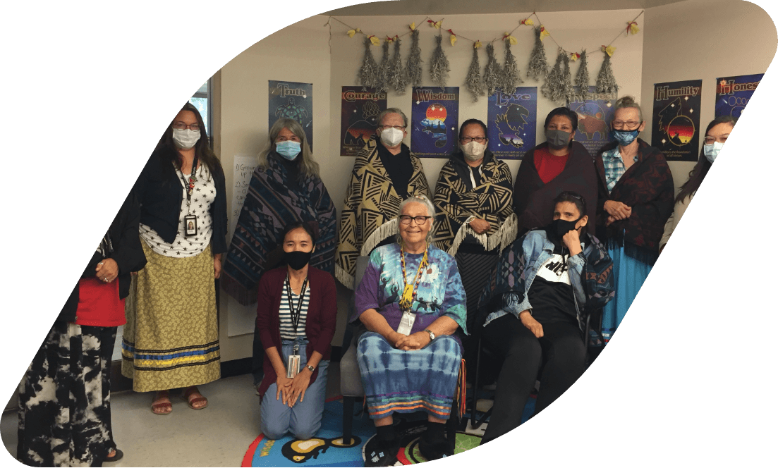 Participants, staff and Elders standing together for a photo. Many are dressed in Indigenous ribbon skirts. Drying sage bundles hang in the background
